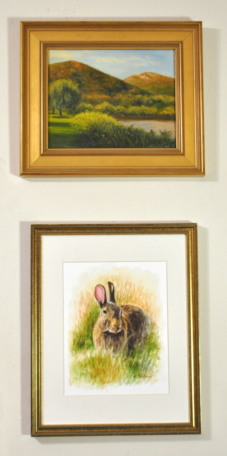 May include: A framed watercolor painting of a brown rabbit sitting in a field of green grass. The rabbit has pink ears and is looking directly at the viewer.