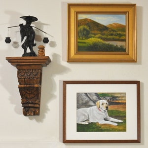 May include: A white framed picture with a painting of a white labrador retriever lying down on a green grassy field. The picture is hanging on a white wall next to a wooden shelf with a black figurine of a person carrying a pole with two baskets on either end.