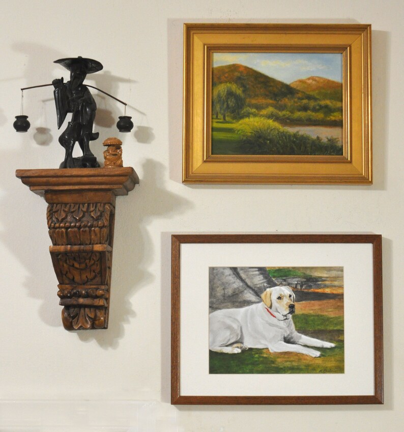 May include: A white framed picture with a painting of a white labrador retriever lying down on a green grassy field. The picture is hanging on a white wall next to a wooden shelf with a black figurine of a person carrying a pole with two baskets on either end.