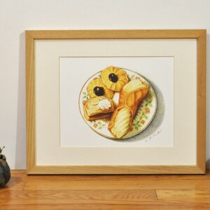 May include: A framed watercolor painting of a plate of pastries. The plate is white with a floral pattern and contains three pastries: a puff pastry, a cannoli, and a cookie. The pastries are all yellow and brown.