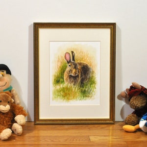 May include: A watercolor painting of a brown rabbit with pink ears sitting in a field of green grass. The painting is framed in a gold frame.