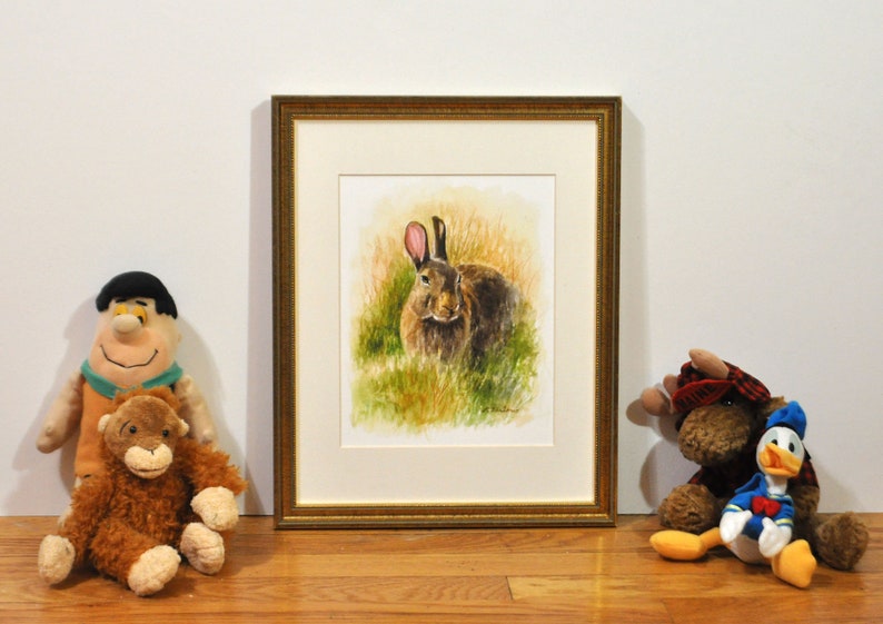 May include: A watercolor painting of a brown rabbit with pink ears sitting in a field of green grass. The painting is framed in a gold frame.