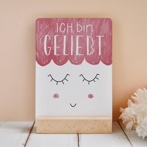 Könnte beinhalten: Ein dekoratives Holzschild mit dem Text "ICH BIN GELIEBT" in weißer Schrift, über einem skurrilen Gesicht mit geschlossenen Wimpern, rosigen Wangen und einem kleinen Lächeln. Das Schild steht auf einem Holzsockel.