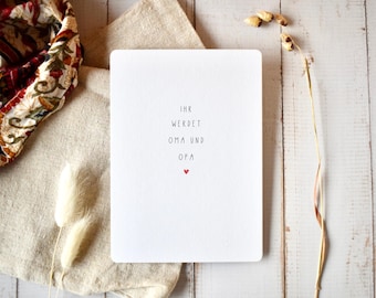 May include: A rectangular white greeting card with rounded corners featuring the black printed text IHR WERDET OMA UND OPA and a small red heart symbol centered below the text. The card is positioned on a light-colored wooden surface.