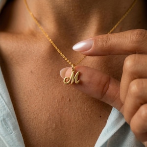 Könnte beinhalten: Eine goldfarbene Halskette mit einer filigranen Kette und einem Anhänger mit dem kursiven Buchstaben "M". Der Anhänger wird von einem Finger gehalten. Die Halskette wird vor einem Hautton-Hintergrund präsentiert.