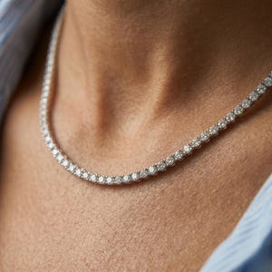 May include: A silver-toned tennis necklace featuring a continuous row of small, round, clear, faceted stones set in a metallic frame. The necklace rests against the skin, showcasing a uniform design of sparkling circular gems linked together.