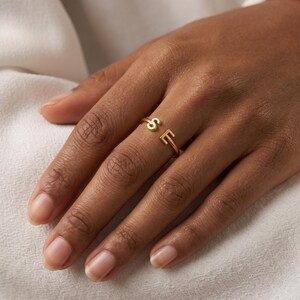 May include: A gold-toned open ring featuring two distinct letters, S and E, positioned at each end of the band. The ring has a polished finish and a simple, clean design that allows for a slight gap between the two capital letters on the finger.