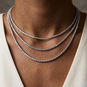May include: Three delicate diamond tennis necklaces of varying lengths are displayed. The necklaces are composed of small, round, sparkling diamonds set closely together. The necklaces are worn on a person's neck, against a white top.