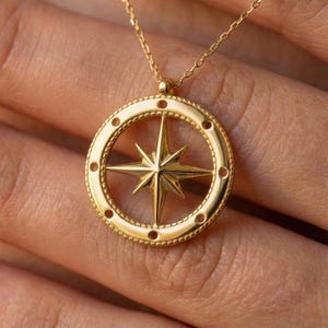 May include: A gold-colored compass pendant necklace. The pendant features a star design within a circular frame with small, evenly spaced holes. The necklace chain is delicate and also gold-colored.