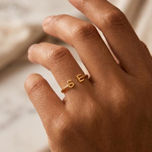 Puede incluir: Un anillo abierto de tono dorado que presenta dos letras mayúsculas, S y E, situadas en los extremos de la banda. El anillo tiene un acabado liso y pulido con un diseño sencillo que permite un ajuste regulable alrededor del dedo.