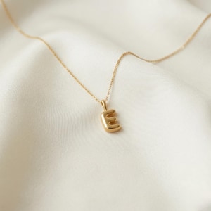 May include: A gold-toned necklace featuring a small, puffed letter E pendant. The pendant is suspended from a delicate, thin gold-toned chain. The design is simple and polished, with a smooth, reflective surface that catches the light clearly.