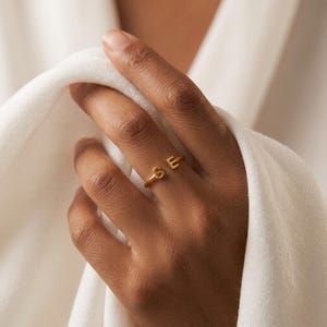 Könnte beinhalten: Ein goldfarbener offener Ring mit zwei Großbuchstaben, S und E, die an den Enden der Ringschiene angebracht sind. Der Ring hat eine glatte, polierte Oberfläche und eine schlichte, dünne Metallkonstruktion, die den Finger teilweise umschließt.