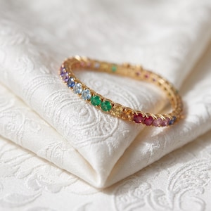Könnte beinhalten: Ein goldenes Tennisarmband mit einer durchgehenden Reihe rund geschliffener Edelsteine in einem lebendigen Regenbogenspektrum, darunter violette, blaue, grüne, gelbe, orangefarbene und rote Steine, gefasst in einen polierten Goldrahmen.