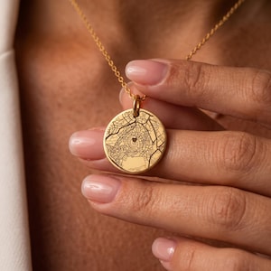 Op de afbeelding: Een goudkleurige ronde hanger aan een ketting met een gedetailleerd, zwart gegraveerd stadsplattegrondontwerp. In het midden van de kaart is een klein hartje zichtbaar. De hanger hangt aan een fijne goudkleurige ketting voor een elegante uitstraling.