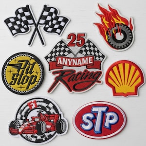 May include: A collection of embroidered racing patches in various designs. Includes checkered flags, a flaming tire, a Shell logo, and patches with text like "Pit Stop", "Anyname Racing", and "STP". The patches feature red, black, white, and yellow colors.