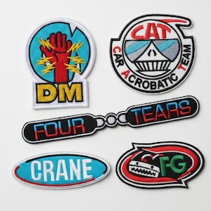 May include: A collection of embroidered patches with various designs and text. One patch features a red hand with yellow lightning bolts and the letters "DM." Another says "CAT CAR ACROBATIC TEAM." Other patches say "FOUR TEARS," "CRANE," and "FG."