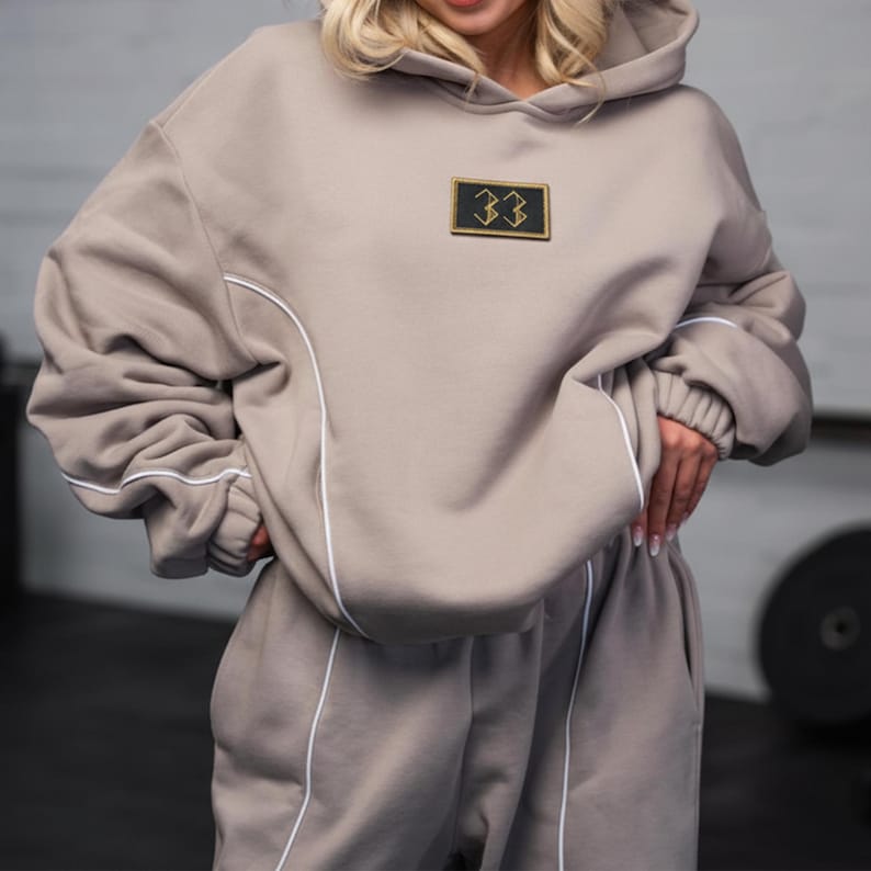 May include: A taupe hoodie and matching sweatpants set. The hoodie features a hood, a patch with the number "33", and white piping along the seams. The sweatpants have white piping along the sides. The outfit is made of a soft, comfortable material.