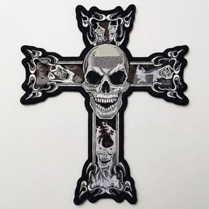 May include: An embroidered patch in the shape of a cross featuring a central human skull with hollow eye sockets and an open jaw. The cross is decorated with intricate flame-like patterns and smaller skull motifs in shades of black, white, and gray.