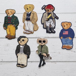 May include: Six embroidered bear patches featuring various outfits: one in a green blazer and yellow pants, one in a tan coat over a USA sweater, one in a polo gear outfit, one in a striped shirt and beret, one in a green jacket, and one in a flag sweater.