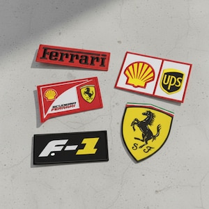 May include: A collection of five patches featuring logos and branding. The patches include the Ferrari logo in red, a Scuderia Ferrari patch, a Shell and UPS patch, a yellow Ferrari shield, and an F-1 patch.