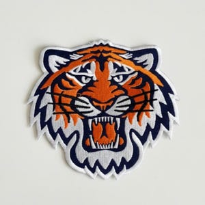May include: Embroidered patch featuring a tiger's head with an open mouth, showcasing sharp teeth. The design uses orange, white, and navy blue thread. The patch has a white border and is likely for application on clothing or accessories.