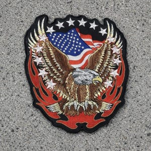May include: An embroidered patch featuring a bald eagle with spread wings centered over a United States flag. The design includes a border of white stars on a black background, accented by red flame-like shapes at the bottom and sides of the eagle.