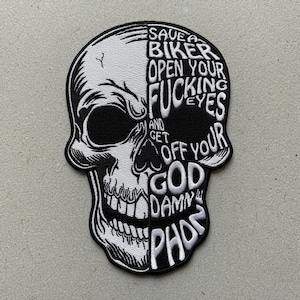 May include: A black and white skull patch with the text "SAVE A BIKER OPEN YOUR FUCKING EYES AND GET OFF YOUR GOD DAMN PHONE". The patch is embroidered with a detailed skull design.