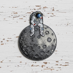 May include: Embroidered patch featuring a dejected astronaut seated on a detailed gray moon. The astronaut is wearing a white spacesuit with red and blue accents. The moon has a textured surface with numerous craters, set against a white, distressed wood background.