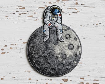 May include: Embroidered patch featuring a dejected astronaut seated on a detailed gray moon. The astronaut is wearing a white spacesuit with red and blue accents. The moon has a textured surface with numerous craters, set against a white, distressed wood background.