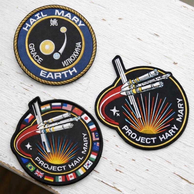 Three embroidered space mission-style patches displayed on a white wooden surface, including a “Hail Mary – Earth” patch with multilingual text and orbital design, and two “Project Hail Mary” patches featuring a spacecraft and radiant burst motif surrounded by international flags, evoking a fictional global space mission theme.