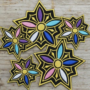 Puede incluir: Parches bordados con forma de flor en varios tamaños. Cada parche presenta un diseño de estrella negra con un borde dorado y pétalos coloridos en tonos morado, rosa, azul y blanco. El centro de cada flor es amarillo.