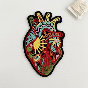 May include: An embroidered patch shaped like an anatomical heart with a black border. The design features vibrant thread colors including red, yellow, light blue, pink, and green, depicting stylized floral, sun, and geometric patterns.