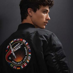 May include: Black bomber jacket featuring a circular patch with a rocket launch design and the text "PROJECT HAIL MARY." The patch is surrounded by flags. The jacket has a ribbed collar and cuffs.