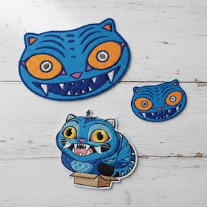 May include: Three blue cat patches with orange eyes and sharp teeth. One patch is a cat head, another is a smaller cat head, and the third is a cat sitting in a box. The patches are embroidered.