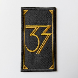 May include: Black rectangular patch with a gold embroidered number "33" in a stylized font. The patch has a gold border and corner accents. The background is black.