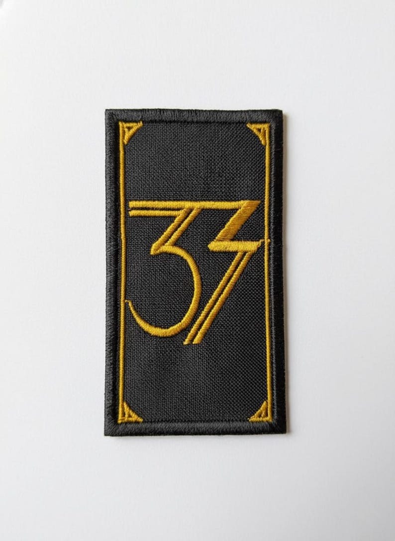 May include: Black rectangular patch with a gold embroidered number "33" in a stylized font. The patch has a gold border and corner accents. The background is black.