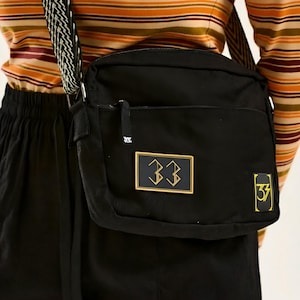 May include: Black crossbody bag with a gold-trimmed rectangular patch featuring the number "33" in gold. The bag has a black and white patterned strap and a small zipper pocket. The bag is made of a durable material.