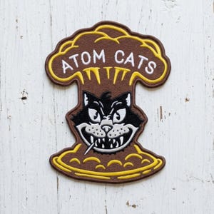 May include: A brown, yellow, and white embroidered patch shaped like a mushroom cloud. The text ATOM CATS is stitched in white at the top. Centered below is a black and white cat face with sharp teeth and a toothpick in its mouth.