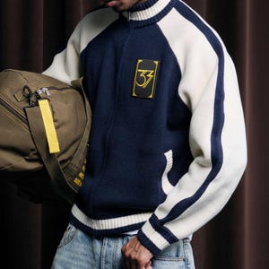 May include: Navy blue and cream-colored varsity jacket with a gold patch featuring the number 33. The jacket has a zip-up front, side pockets, and white stripes down the sleeves. A green duffel bag with a yellow tag is also visible.