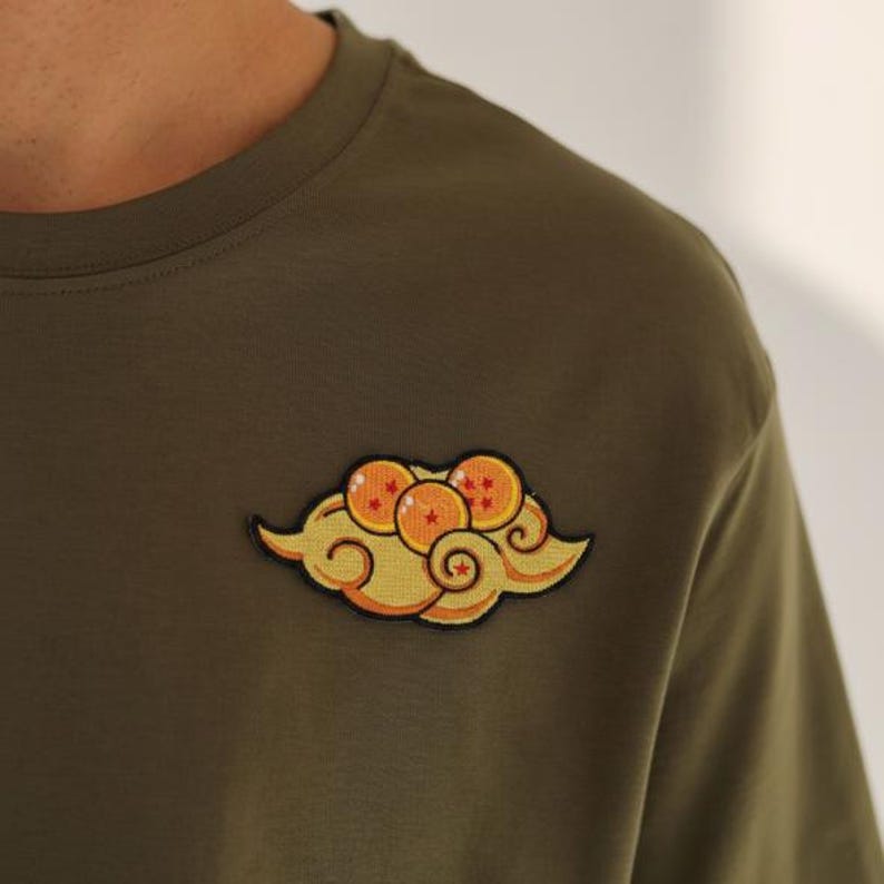 May include: Olive green t-shirt with a yellow and orange cloud patch featuring orange spheres with stars. The patch is on the left side of the shirt, near the shoulder.