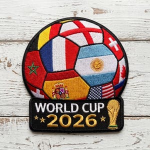 May include: A circular embroidered patch featuring a soccer ball design composed of various national flags, including Morocco, France, Argentina, Spain, and the USA. Below the ball, a black rectangular base displays the white text WORLD CUP 2026.
