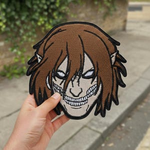 May include: Embroidered patch featuring a brown-haired character with a wide, toothy grin and intense white eyes. The patch has a black border and is likely for application on clothing or accessories. The design is detailed and stylized.