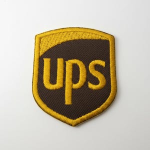 May include: A brown and gold UPS patch. The patch is shield-shaped with a brown background and gold embroidered border. The letters "UPS" are embroidered in gold.