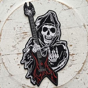 May include: An embroidered patch featuring a skeleton in a black hooded robe holding a black electric guitar with red flame details. The skeleton has one hand on the guitar neck and the other hand raised in a rock and roll gesture.