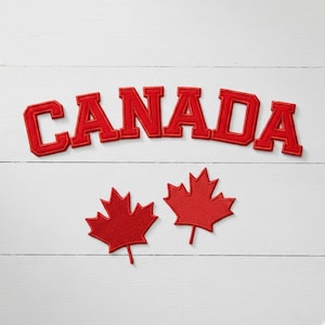 May include: Red felt applique spelling "CANADA" above two red maple leaf appliques on a white wooden background. The letters are in a bold, collegiate style. The maple leaves are a symbol of Canada.