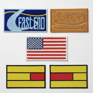 May include: A collection of embroidered patches. One patch features the text "EAST END" with a wave design. Another displays "Frontier" in a script font. An American flag patch is also present, along with two patches with yellow and red geometric designs.