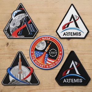 May include: A collection of embroidered NASA and Artemis mission patches. The patches feature rockets, the moon, and the NASA logo. Some patches are triangular, while others are circular. The text includes "ARTEMIS" and "THE ROCKET'S RED GLARE."