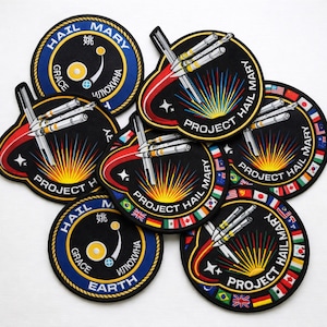 May include: A collection of embroidered patches with a space theme. The patches feature rockets, the words "Project Hail Mary," and other text in multiple languages. The patches have a black background with colorful accents and borders.