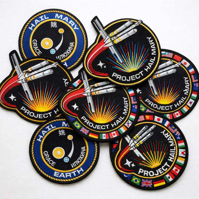 May include: A collection of embroidered patches with a space theme. The patches feature rockets, the words "Project Hail Mary," and other text in multiple languages. The patches have a black background with colorful accents and borders.