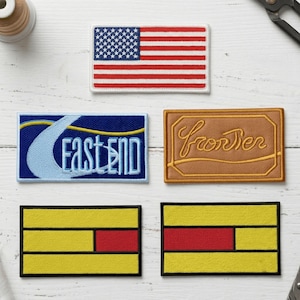 May include: Five rectangular embroidered patches. Top: a United States flag. Middle left: a blue patch with the text "EAST END" in white and a yellow wave. Middle right: a tan patch with the word "frontier" in cursive. Bottom: two yellow patches with red blocks.